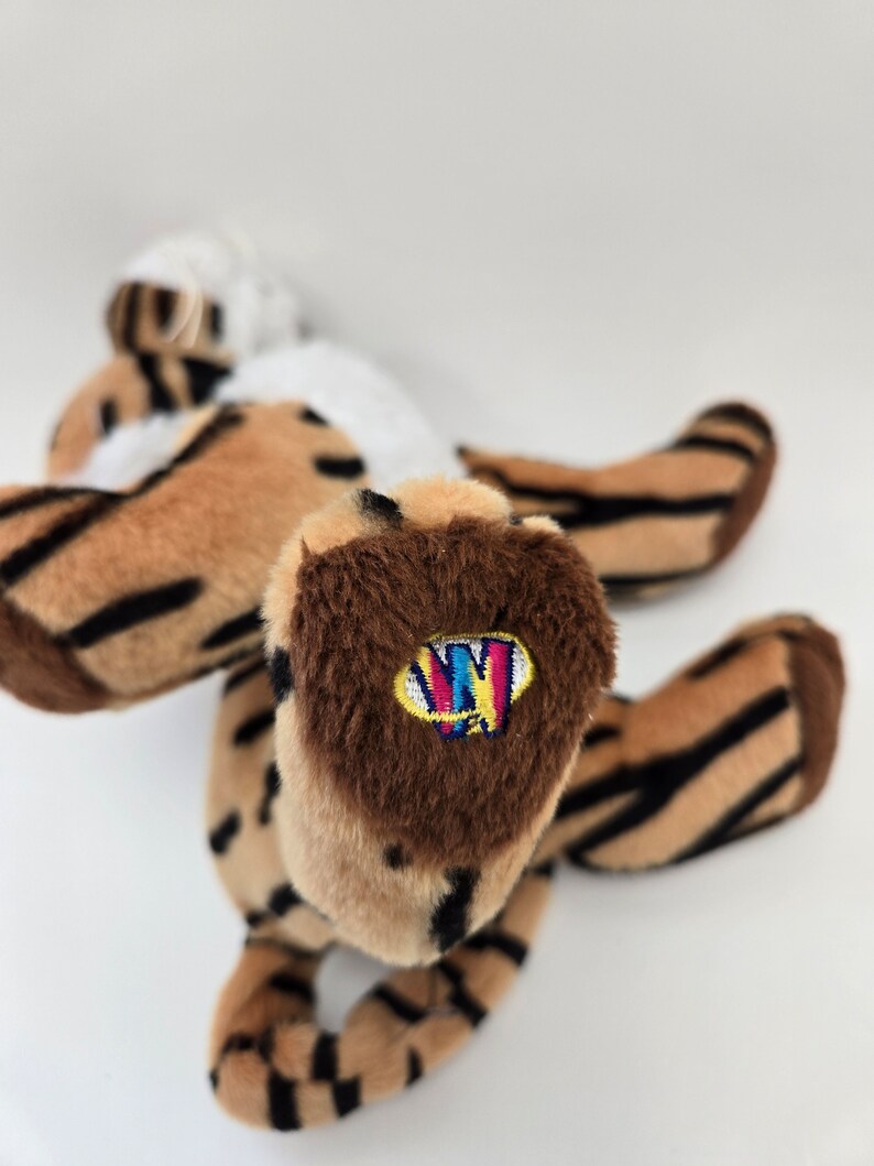 Puede incluir: Un tigre de peluche rayado marr&oacute;n y negro con vientre blanco. El tigre tiene una pata marr&oacute;n con un logotipo "Ty" colorido en ella.