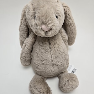 May include: A plush, light brown bunny with long, floppy ears, a pink nose, and a soft, textured appearance. The stuffed animal has a smiling expression and is designed for children.