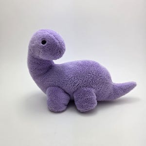 May include: A plush, lavender-colored dinosaur toy with a long neck and small, stubby legs. The stuffed animal has a soft, fuzzy texture and a small, round eye. The dinosaur's tail is short and pointed, and it is set against a white background.