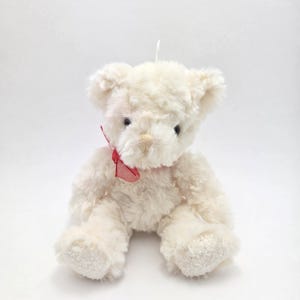Puede incluir: Un oso de peluche blanco con un lazo rojo sentado sobre un fondo blanco.