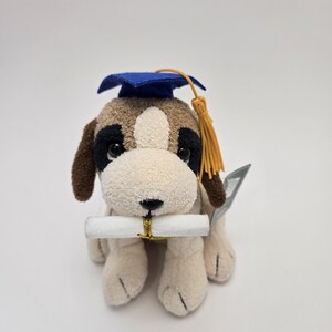 May include: A plush dog wearing a blue graduation cap and gown, holding a white scroll in its mouth. The dog has brown and white fur and a gold tassel on its cap.