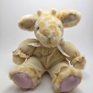 May include: A plush giraffe toy with a soft, yellow and white spotted head and ears. The giraffe wears a light yellow onesie with ruffled sleeves and a white ribbon with yellow polka dots. The feet are pink with a textured pattern.