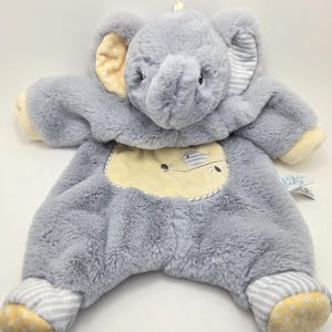 Peut inclure: Un jouet en peluche gris en forme d'éléphant pour bébé avec des accents jaunes et blancs. L'éléphant porte une tenue rayée gris et blanc avec un patch jaune sur la poitrine. Le jouet a une boucle sur le dessus de la tête pour l'accrocher.