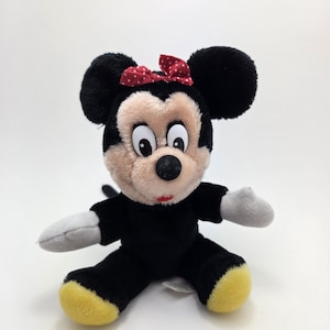Op de afbeelding: Een pluche Minnie Mouse pop. De pop heeft zwarte vacht, grote zwarte oren en een rode strik met witte stippen. Het gezicht is beige, de handschoenen wit en de voeten geel. Een klassiek Disney-personage.