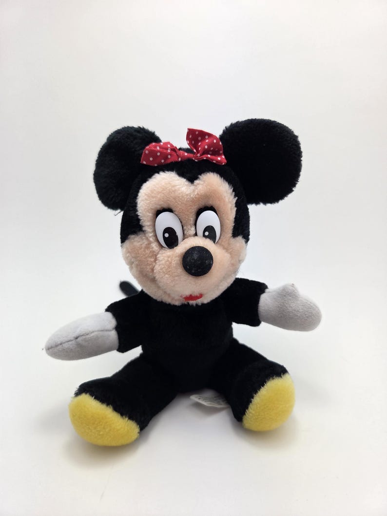 Op de afbeelding: Een pluche Minnie Mouse pop. De pop heeft zwarte vacht, grote zwarte oren en een rode strik met witte stippen. Het gezicht is beige, de handschoenen wit en de voeten geel. Een klassiek Disney-personage.