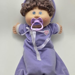 Peut inclure: Poupée Cabbage Patch Kids vêtue d'une tenue lavande et d'un bonnet assorti. La poupée a des cheveux bruns, des yeux bleus et une sucette. La tenue a un ruban blanc et un écusson en forme de cœur avec "CPK".