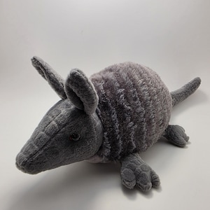 May include: A plush, gray armadillo toy with a textured, segmented body and a long tail. The stuffed animal has small, rounded ears and four tiny feet. The toy is made of soft, fuzzy material.