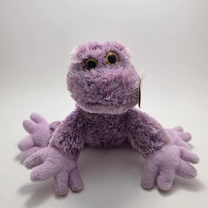 Peut inclure: Une peluche en forme de grenouille violette avec de grands yeux dorés. La grenouille a une fourrure douce et floue et des bras et des jambes tendus. Ce jouet est conçu pour ressembler à une grenouille et convient aux enfants.