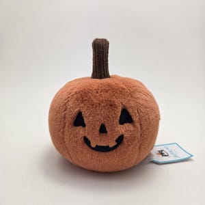 May include: A plush, orange pumpkin with a smiling jack-o'-lantern face and a brown stem. The soft toy is designed for Halloween and features embroidered black eyes and a mouth. A small tag is attached.