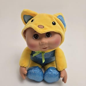 Peut inclure: Un jouet en peluche jaune portant un costume d'animal à capuche bleu et vert. Le jouet a un visage souriant et est assis les jambes croisées.