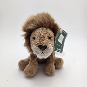 Puede incluir: Un león de peluche con melena, cuerpo y patas marrones. El león tiene una cara marrón claro y una nariz negra. Una etiqueta está adjunta. El juguete está diseñado para parecer un león amigable.