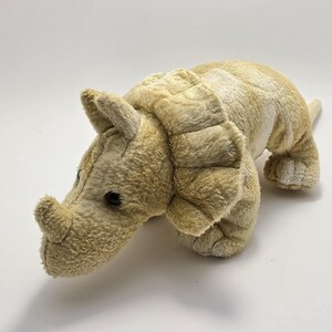 May include: A plush triceratops dinosaur toy featuring soft, light tan fur, a prominent frill around the neck, a single horn on the snout, and two smaller horns above the eyes. The stuffed animal has dark, round eyes and a textured, fuzzy surface.