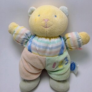 May include: A plush teddy bear with a yellow head, pastel striped sleeves, and overalls featuring a peach-colored leg and a light green leg. The green leg has the embroidered numbers 123 in pink, yellow, and blue. A blue Eden brand tag is attached.