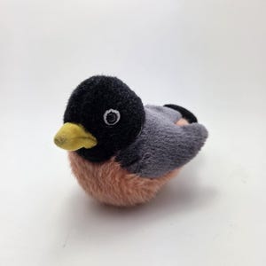 Può includere: Un giocattolo a forma di uccello di peluche con testa nera, ali grigie e petto color pesca. L'uccello ha un becco giallo e occhi bianchi. Il giocattolo è progettato per assomigliare a un pettirosso.