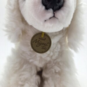 May include: A plush white dog with a black nose and eyes. The dog has soft, fluffy fur and a round, brown tag with the words "Aurora Miyoni Tots". The dog is a stuffed animal.