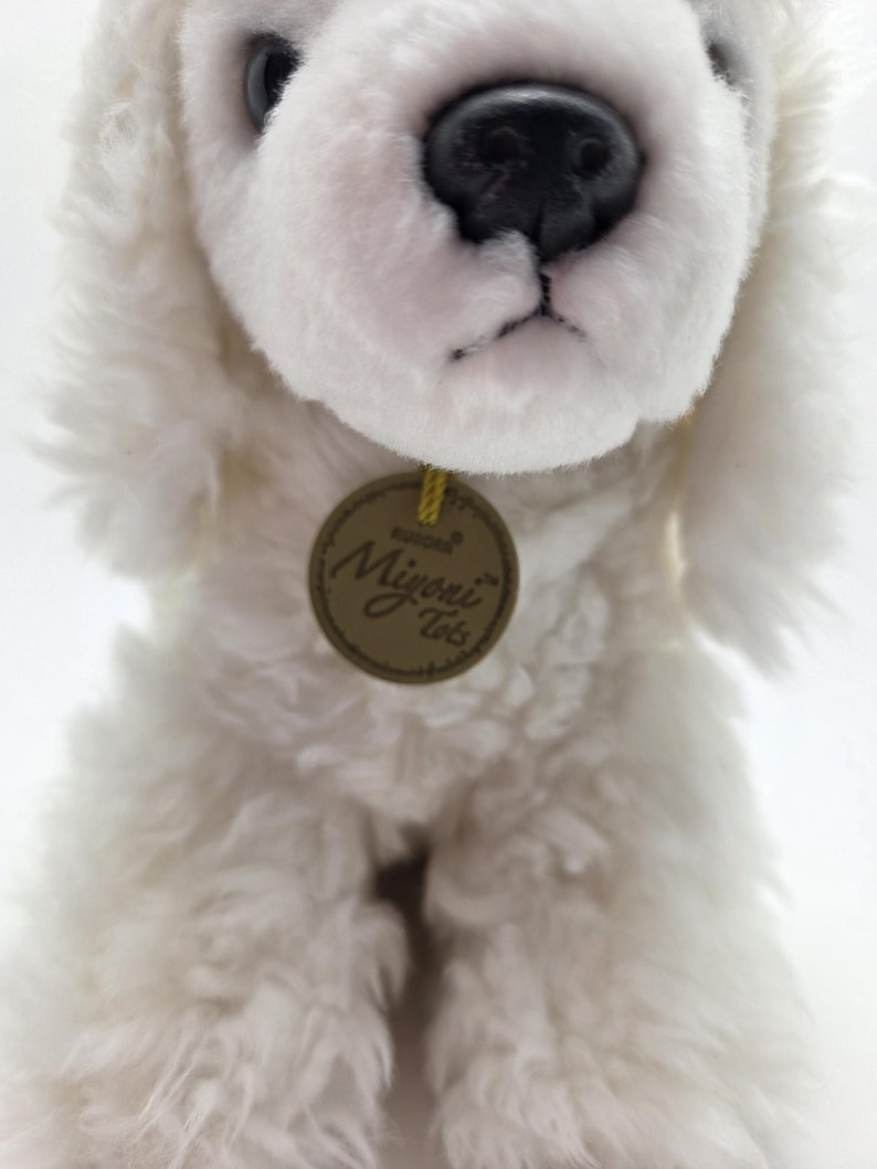 May include: A plush white dog with a black nose and eyes. The dog has soft, fluffy fur and a round, brown tag with the words "Aurora Miyoni Tots". The dog is a stuffed animal.