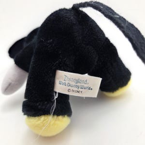 Op de afbeelding: Een zwart pluche speelgoed met gele voeten en een wit oor. Het speelgoed heeft een label met de tekst "Disneyland Walt Disney World &copy; DISNEY". Het speelgoed is gemaakt van een zacht, pluizig materiaal.