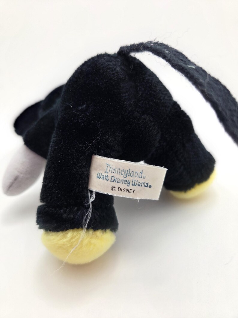 Op de afbeelding: Een zwart pluche speelgoed met gele voeten en een wit oor. Het speelgoed heeft een label met de tekst "Disneyland Walt Disney World &copy; DISNEY". Het speelgoed is gemaakt van een zacht, pluizig materiaal.