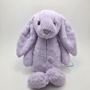 Puede incluir: Un conejo de peluche lavanda con orejas largas y caídas y un aspecto suave y texturizado. El peluche tiene una nariz de color púrpura claro y está sentado erguido. Los pies del conejo son redondeados y esponjosos. Una etiqueta está adjunta al conejo.