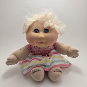 May include: A Cabbage Patch Kids doll with blonde hair and blue eyes. The doll is wearing a pink top with a floral pattern and a striped skirt with pink, yellow, and blue stripes.