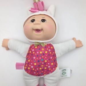 May include: A Cabbage Patch Kids doll dressed as a unicorn. The doll has a white hooded outfit with a pink and polka-dotted bodice, pink shoes, and a gold horn. The doll's arms are outstretched.