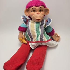Op de afbeelding: Een pluche apenpop met een felroze pluizige muts, een vinyl gezicht en rode stoffen benen. De pop draagt een gestreept schort met groene, witte, beige en donkerblauwe verticale lijnen over groenblauwe mouwen. In één hand wordt een kleine gele plastic banaan vastgehouden.