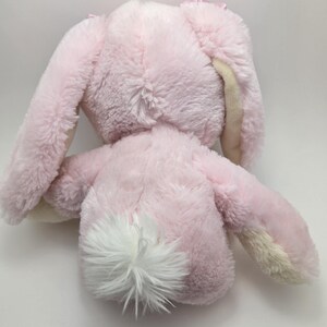 May include: A plush pink bunny toy with long ears and a fluffy white tail. The bunny's fur is soft and textured, with a light pink color and cream-colored accents on the ears. The toy is designed for children.