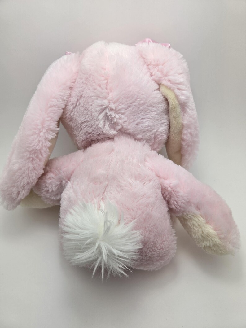 May include: A plush pink bunny toy with long ears and a fluffy white tail. The bunny's fur is soft and textured, with a light pink color and cream-colored accents on the ears. The toy is designed for children.