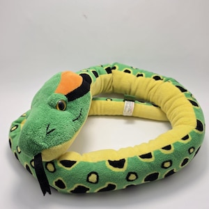 May include: A plush snake toy featuring a green body with black spots and a yellow underside. The head has an orange patch on top, yellow eyes, and a black felt tongue protruding from the mouth. A small white fabric tag is attached to the side.