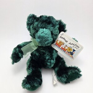 May include: A plush, emerald green teddy bear with a matching ribbon tied around its neck. The bear has a tag that reads "Animal Junction by Russ" with a train illustration. The bear is made of soft, fuzzy material.