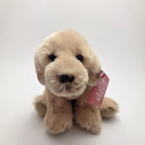 Pode incluir: Um cachorrinho de pelúcia golden retriever em tom castanho claro. O brinquedo de pelúcia tem olhos pretos e um nariz preto. Uma etiqueta rosa está presa à orelha.