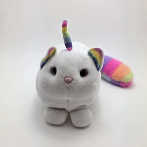 Puede incluir: Un peluche blanco con forma de gato con un cuerno y una cola de unicornio arcoíris. El gato tiene ojos negros, una nariz rosa y detalles de arcoíris en las orejas. La cola está rayada con varios colores.