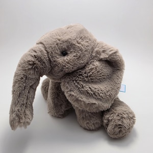 May include: A plush elephant toy made of soft, light brown faux fur. The stuffed animal features a long, curved trunk, large floppy ears, and small black bead eyes. A small blue tag is visible on the side of the elephant's body.