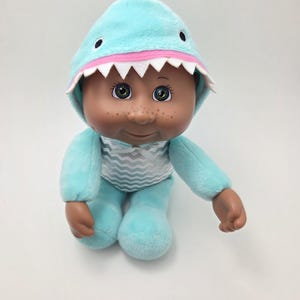 May include: A plush doll wearing a light blue shark costume with a matching hood. The hood has white jagged teeth and black eyes. The doll has brown skin, freckles, and green eyes. The doll's outfit has a white and gray striped pattern.