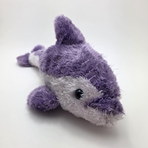 May include: A plush toy dolphin in shades of purple and white. The dolphin has a soft, fuzzy texture, with a prominent dorsal fin and flippers. It features a black eye and a smiling expression, designed for children.