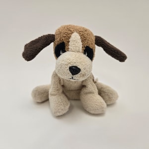 Może przedstawiać: Pluszowa zabawka w kształcie psa z brązowo-białą głową, brązowymi uszami i czarnym nosem. Zabawka siedzi na białej powierzchni. Pies ma czarne oczy i biały pasek na twarzy.