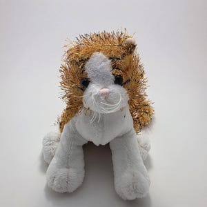 May include: A plush cat toy featuring white fur on the face, chest, and paws, contrasted with a textured, shaggy orange and black striped pattern on the head and back. The toy has dark eyes, a small pink nose, and white whiskers.