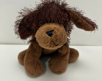 Op de afbeelding: Een bruin pluchen hondenspeelgoed met een harige kop en flaporen. De hond heeft een lichtbruin gezicht en poten, met een zwarte neus en ogen. Het speelgoed staat op een witte ondergrond.