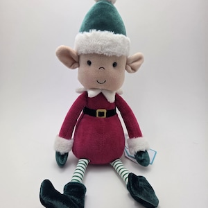 May include: A soft elf doll featuring a green hat with white trim and a white pom-pom. The doll wears a red outfit with a black belt, green and white striped legs, and green shoes. The elf has a smiling face and large ears.