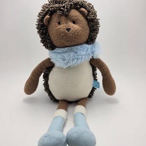 Puede incluir: Un peluche de erizo con una cabeza espinosa marrón, brazos marrones y piernas azul claro. El erizo tiene un cuerpo blanco y una bufanda azul claro. El juguete está diseñado para ser suave y tierno.