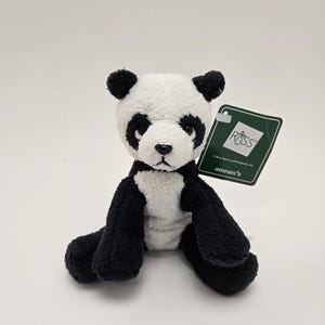 May include: A small black and white plush panda bear sitting on a white surface. The panda has black ears, eyes, and paws. The panda is wearing a tag that says "Russ" and "amram's".
