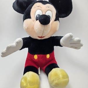 May include: A plush Mickey Mouse toy. The toy has a black body, ears, and head, with a tan face and white gloves. It wears red shorts with yellow buttons and yellow shoes. The toy's arms are outstretched.