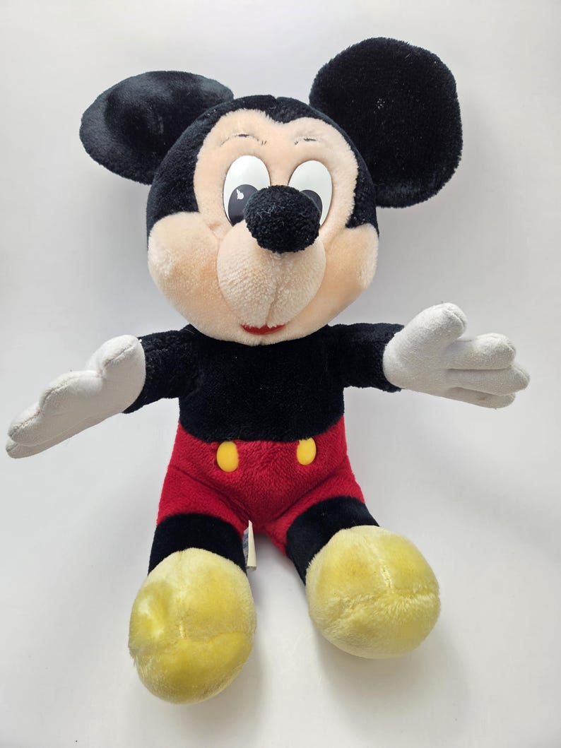 May include: A plush Mickey Mouse toy. The toy has a black body, ears, and head, with a tan face and white gloves. It wears red shorts with yellow buttons and yellow shoes. The toy's arms are outstretched.