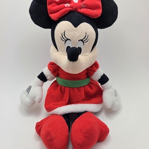 May include: A plush red and white Christmas Minnie Mouse toy with a green belt and a red bow with white snowflakes.
