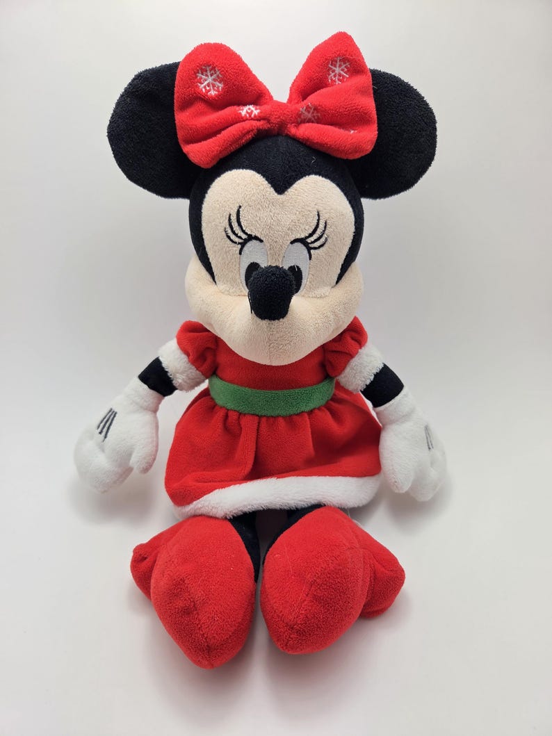 May include: A plush red and white Christmas Minnie Mouse toy with a green belt and a red bow with white snowflakes.