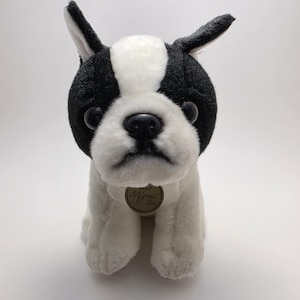 May include: A plush Boston Terrier dog toy with black and white fur. The toy has a black nose, large black eyes, and a small gold tag. The dog is sitting upright with its front paws extended.
