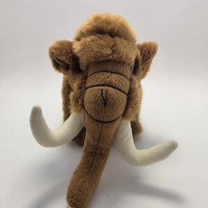 May include: A plush woolly mammoth toy featuring soft brown fur, two large curved off-white tusks, and a prominent trunk. The creature has small ears, dark eyes, and stitched detailing on the face, creating a realistic representation of the prehistoric animal.