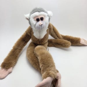 Può includere: Una scimmia di peluche con corpo marrone, testa grigia e viso bianco. Il giocattolo ha braccia e gambe lunghe con mani e piedi rosa. La scimmia è seduta su una superficie bianca.