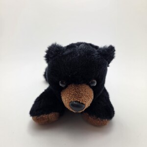 May include: A plush black bear cub toy with a brown snout and paws. The bear has small, dark eyes and is lying down on a white surface. The toy is made of soft, fuzzy material.