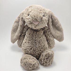 May include: A plush stuffed animal in the shape of a rabbit with long, floppy ears. The toy is covered in textured, shaggy, light beige fur and features a small, smooth, pale pink nose centred on its face. It is seated in an upright position.